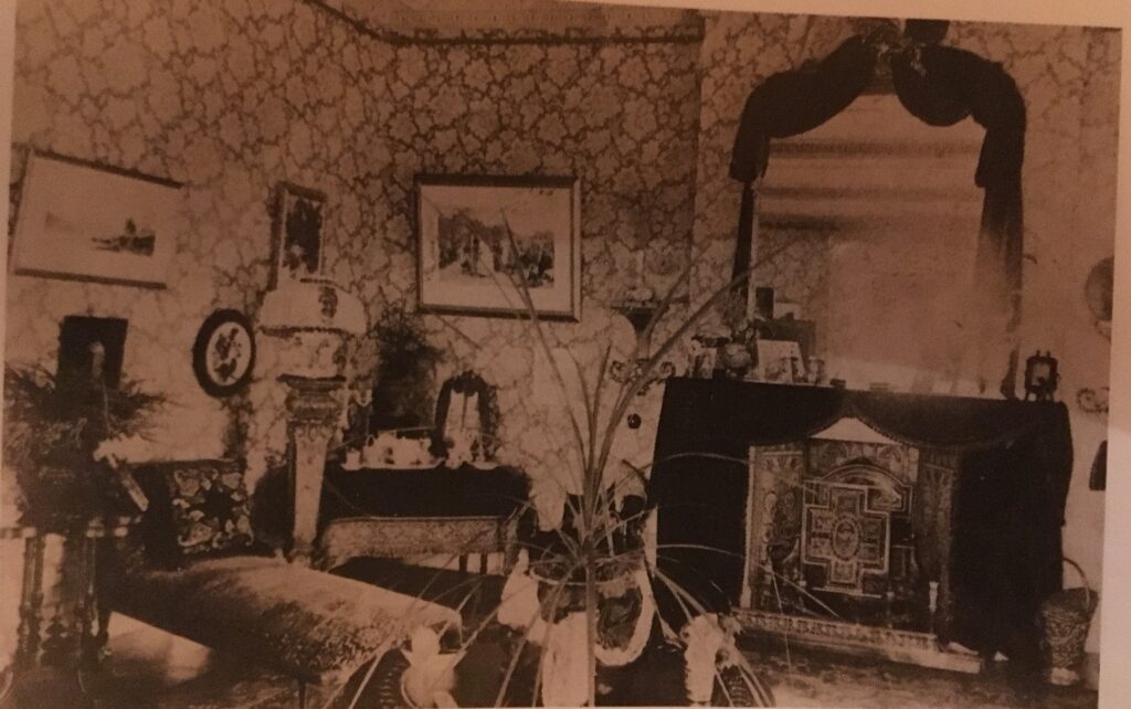 Black and white photograph of middle class parlour showing typical Victorian interior style with a fireplace and mantle mirror draped with pelmet and curtains.